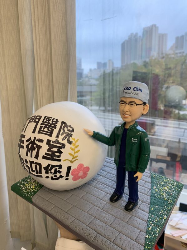 穿手術帽與深藍手術服的麻醉科醫生主題人像公仔，外搭綠色外套，右手指向一個寫有「屯門醫院 手術室 歡迎您！」的大型白色球形展示物；底座為灰色磚路與綠色草地點綴，背景是窗外高樓天際線。適合作為醫護致謝或退休紀念禮物。