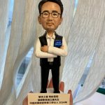 手掌托著一尊男性警察人像公仔：短髮、配眼鏡與淡鬍渣，白色襯衫外穿黑色背心，雙臂交疊，左臂夾著藍色小冊（印有 POLICE CRM CDIST 字樣），黑色長褲與皮鞋。站在白色圓形底座，前方名牌印有祝賀文案與年份。背景為室內窗簾與桌面，整體呈現榮休致敬的送禮氛圍。