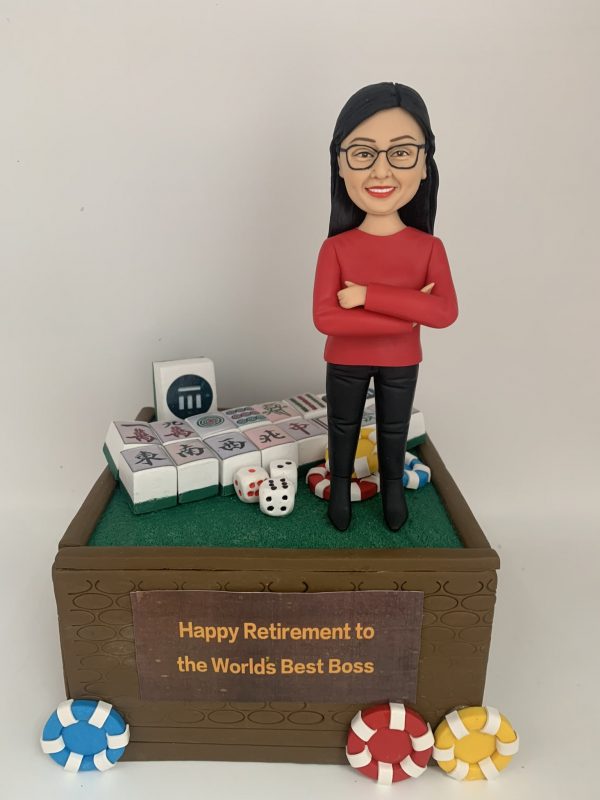 專屬訂製的人像公仔，以女性上司為原型，身穿紅色上衣與黑色長褲，雙手交叉微笑站立。底座設計成麻將桌風格，擺放多張麻將牌、骰子與籌碼，前方木製牌匾寫有「Happy Retirement to the World's Best Boss」，象徵榮退賀禮與專屬紀念。此人像公仔將 麻將元素 與感謝心意融合，打造獨特的退休祝福禮物。
