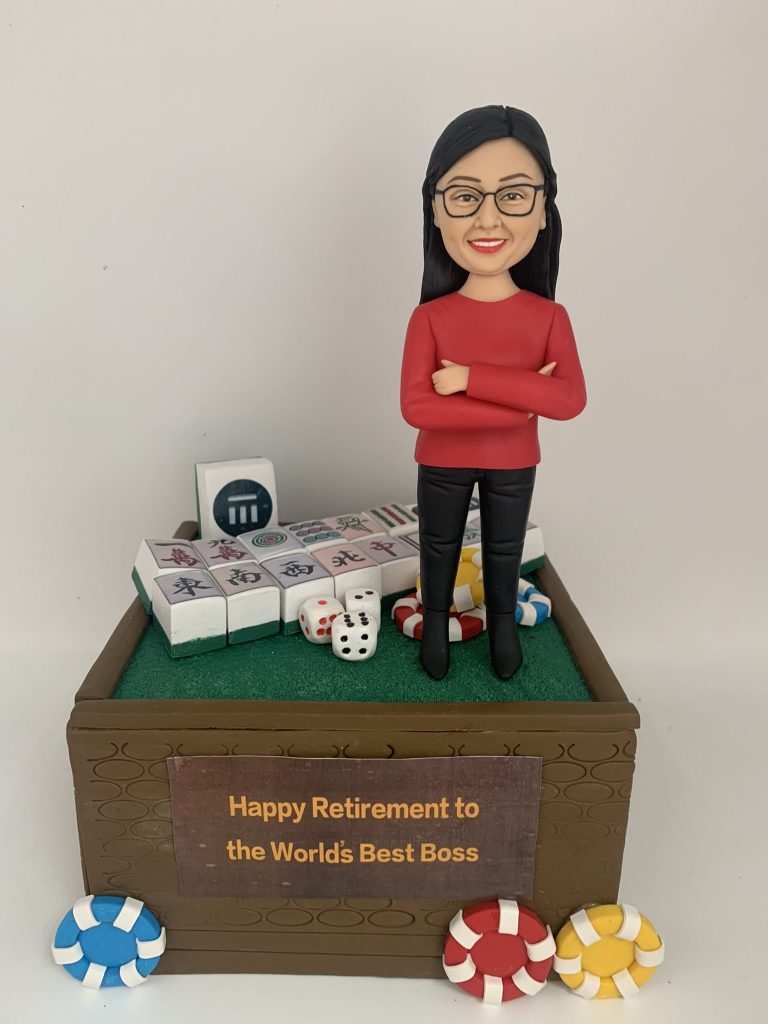 專屬訂製的人像公仔，以女性上司為原型，身穿紅色上衣與黑色長褲，雙手交叉微笑站立。底座設計成麻將桌風格，擺放多張麻將牌、骰子與籌碼，前方木製牌匾寫有「Happy Retirement to the World's Best Boss」，象徵榮退賀禮與專屬紀念。此人像公仔將 麻將元素 與感謝心意融合，打造獨特的退休祝福禮物。