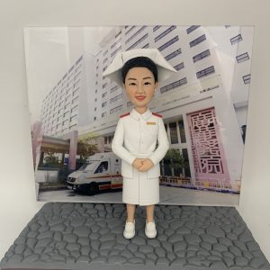 訂製醫護人像公仔，身穿白色護士制服與傳統護士帽，背景為醫院大樓與停泊的救護車，展示專業護士的典雅形象。這款公仔象徵醫生與醫護人員多年服務的感謝與榮耀，特別適合作為退休、升遷或致敬禮物。