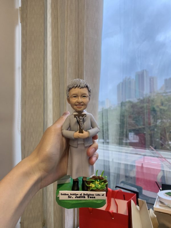 一款手持的客製化修女人像公仔，為慶祝其獻身生活50週年金慶而設，公仔手持十字架，底座刻有英文紀念字句，是送給修女或牧師等神職人員的人像公仔禮物。
