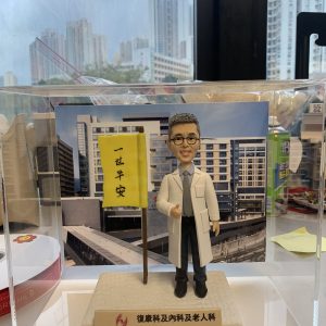 訂製醫生人像公仔，身穿白袍、佩戴眼鏡，手持寫有「一世平安」旗幟，底座標示「屯門醫院」，背景為醫院大樓。此公仔專屬於復康科、內科及老人科醫生，象徵醫護專業精神與祝福心意，適合作為醫生感謝禮物或退休紀念品。