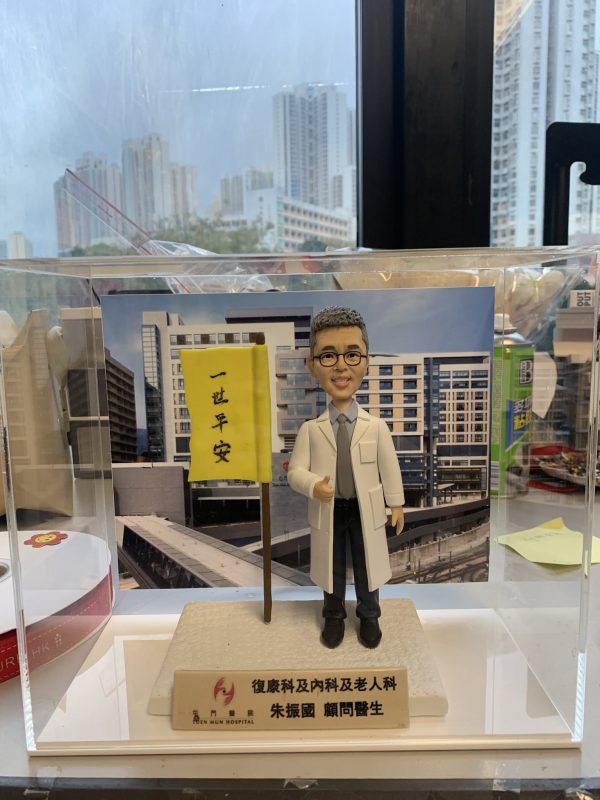 訂製醫生人像公仔，身穿白袍、佩戴眼鏡，手持寫有「一世平安」旗幟，底座標示「屯門醫院」，背景為醫院大樓。此公仔專屬於復康科、內科及老人科醫生，象徵醫護專業精神與祝福心意，適合作為醫生感謝禮物或退休紀念品。