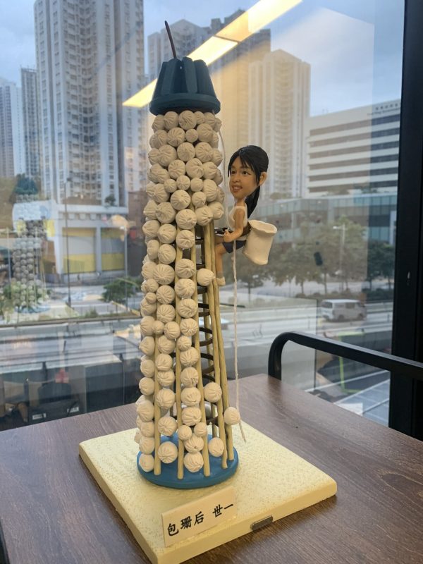 一款搶包山主題的人像公仔場景模型：中心是一座覆滿白色小包的圓柱形包山塔，頂部藍色帽狀結構，側邊設有竹梯與垂繩；一名留黑色馬尾的女性造型，著運動背心與短褲，腰間掛白色收集袋，雙手抓梯向上攀爬，表情專注帶笑。