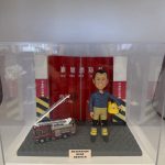 一款為高級消防隊長林龍坤訂製的退休消防人像公仔，公仔身穿制服，與一輛升降台消防車模型一同站立在油塘消防局紅色鐵閘背景前，底座刻有油塘消防局C隊的榮休祝福。