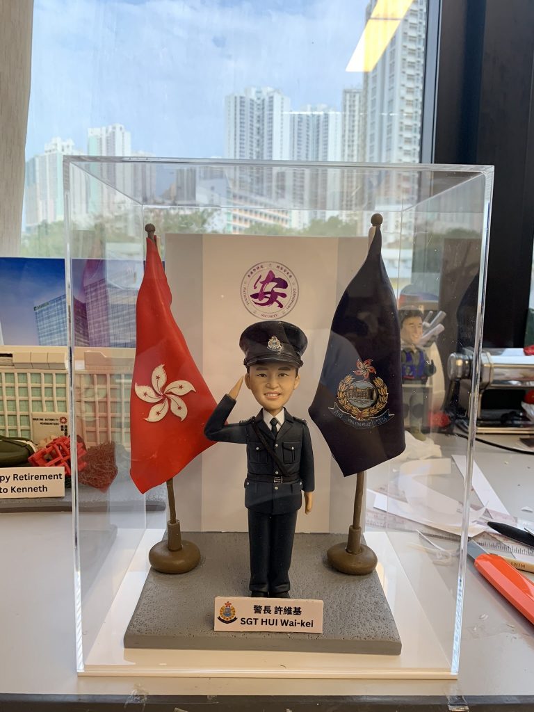 透明展示盒內的警察人像公仔：身著深藍制服與制帽，右手舉至眉間敬禮，左手自然下垂；胸前有肩帶與口袋釦飾，黑色皮鞋。人物站在灰色方形地台中央，左右各有一面旗幟：左側為紅底白花旗，右側為深藍色警徽旗。前方白底名牌印有中英對照的職級稱呼與姓名；名牌左側帶有警徽圖案。背板中央見到圓形紫色標誌，展示盒置於工作桌上，周圍有手工具與模型配件；窗外為高樓景觀。整體呈現正式、榮譽與頒贈性質，適合作為升遷、榮休或部門致意的送禮。