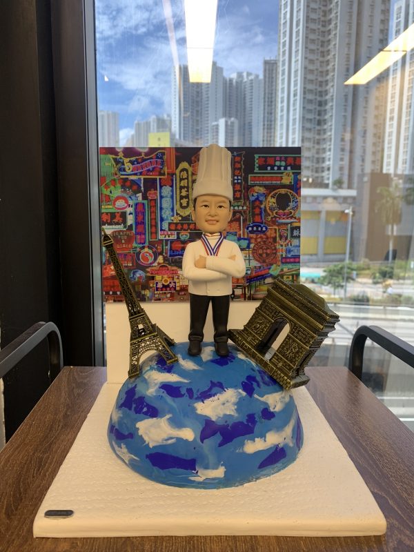 一款廚師人像公仔站在地球模型上，象徵環遊世界，是送給廚師的完美退休禮物，公仔旁有巴黎地標，背景為香港霓虹燈。