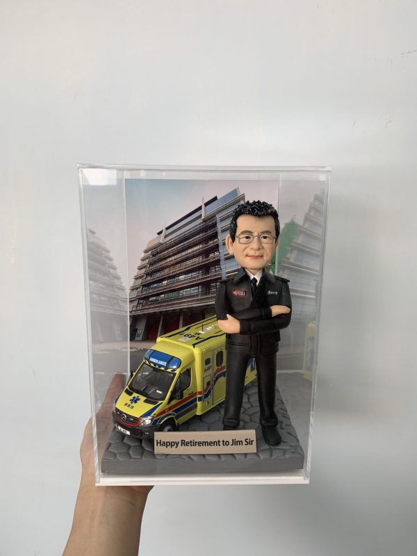 一款為救護員長官 Jim Sir 訂製的退休消防人像公仔，公仔身穿制服，與一輛香港救護車模型一同站立在消防局照片背景前，底座刻有退休祝福語。