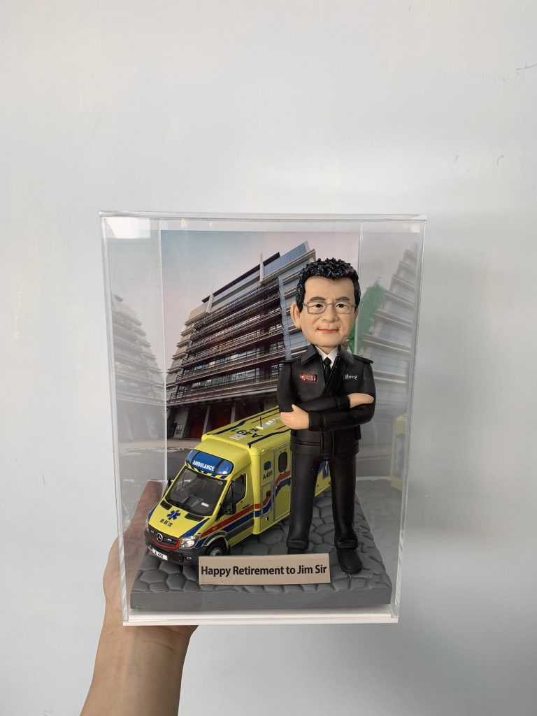 一款為救護員長官 Jim Sir 訂製的退休消防人像公仔，公仔身穿制服，與一輛香港救護車模型一同站立在消防局照片背景前，底座刻有退休祝福語。