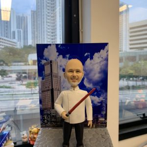酒店主廚主題人像公仔：光頭留灰白山羊鬍的男廚師穿白色雙排釦主廚外套、黑長褲與黑鞋，右手握紅黑色長棒狀道具微笑站立。公仔置於灰色石磚紋底座，背板印有藍天白雲與高樓大廈的城市酒店天際線。背景為辦公室窗邊與高樓景色，整體具有都會質感與紀念氛圍，適合作為酒店廚師退休禮物。
