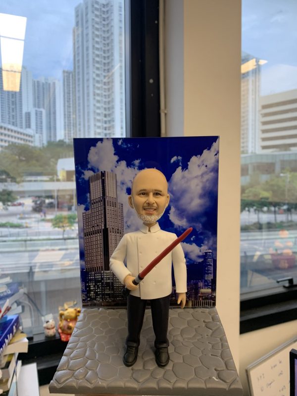 酒店主廚主題人像公仔：光頭留灰白山羊鬍的男廚師穿白色雙排釦主廚外套、黑長褲與黑鞋，右手握紅黑色長棒狀道具微笑站立。公仔置於灰色石磚紋底座，背板印有藍天白雲與高樓大廈的城市酒店天際線。背景為辦公室窗邊與高樓景色，整體具有都會質感與紀念氛圍，適合作為酒店廚師退休禮物。