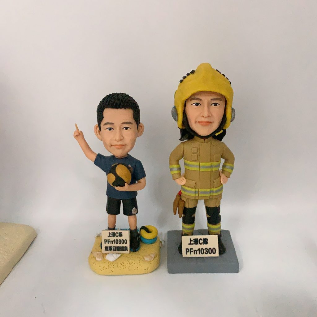 一款為消防員訂製的退休人像公仔禮物，包含兩個公仔，一個穿消防制服，另一個穿休閒服，背景是上環消防局照片，裝在透明保護盒中。
