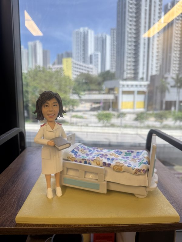 一款為護士訂製的退休禮物。精緻的人像公仔身穿白色護士服，手持記錄簿，面帶微笑地站在一張鋪有花朵圖案被子的醫院病床旁邊，是一份完美還原其工作場景、充滿敬意的紀念品。