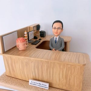 神經外科醫生人像公仔，身穿西裝的男醫生坐在診間辦公桌後方，桌面擺放大腦模型與脊椎模型，象徵專業的神經外科醫學。辦公桌上有電腦、鍵盤、醫學書籍與醫療文件，細緻還原醫生診間場景。此客製化醫護公仔適合作為醫生禮物、醫護紀念品、退休紀念或升職賀禮。