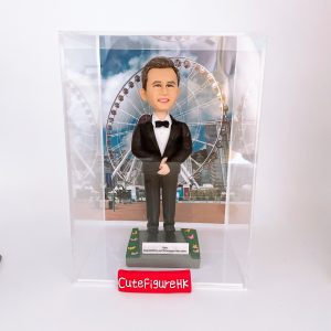 一款為外國同事訂製的人像公仔禮物，公仔是一位穿著燕尾服的男士，站立在以香港摩天輪為背景的場景中，並配有刻上祝福語的底座和透明保護盒。