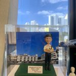 透明展示盒內的警察人像公仔站於綠色草地質感台座：短髮、白色短袖制服襯衫、深色長褲與黑色長筒靴，頭戴深色貝雷帽，雙臂交疊抱著頭盔；腰間配有裝備腰帶。公仔前方金屬名牌印有多行文字，提及機動部隊與PTU等字樣，以及致贈單位資訊。背板為一幅在藍天白雲下的大型白色建築群照片，與公仔台座連成一體。展示盒置於室內架上，四周可見其他模型與擺設，窗外高樓天際線清晰。