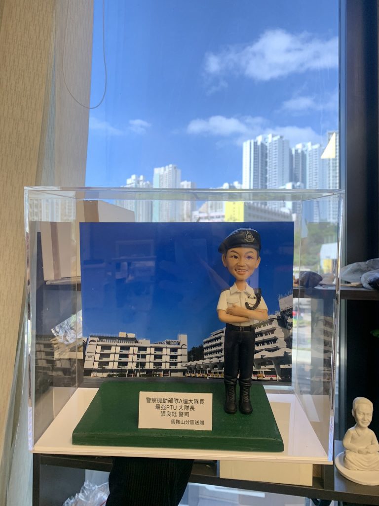 透明展示盒內的警察人像公仔站於綠色草地質感台座：短髮、白色短袖制服襯衫、深色長褲與黑色長筒靴，頭戴深色貝雷帽，雙臂交疊抱著頭盔；腰間配有裝備腰帶。公仔前方金屬名牌印有多行文字，提及機動部隊與PTU等字樣，以及致贈單位資訊。背板為一幅在藍天白雲下的大型白色建築群照片，與公仔台座連成一體。展示盒置於室內架上，四周可見其他模型與擺設，窗外高樓天際線清晰。