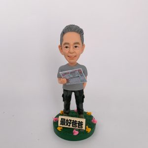 一款栩栩如生的人像公仔，呈現一位慈祥父親的造型，手中拿著帶有賭馬資訊的報紙，展現日常生活興趣。公仔底座上有「最好爸爸」字樣，並以彩色小花裝飾，傳遞濃厚的家庭溫馨氛圍與祝福。這是一份結合專屬興趣與情感的特別訂製人像公仔。
