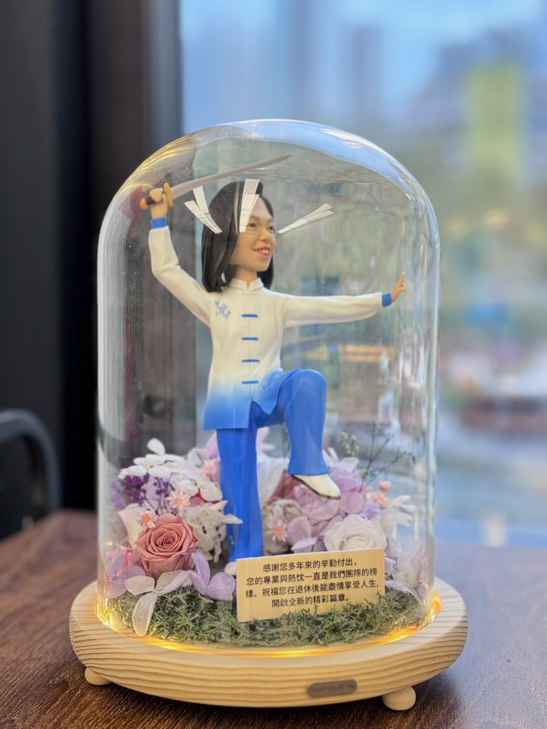 太極人像公仔展示於玻璃永生花燈罩內,公仔身穿藍白太極服,手持太極劍,單腳提腿擺出太極動作,底座裝飾有粉紫玫瑰與永生花,並附上感謝與祝福字句,作為退休禮物擺設。
