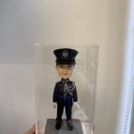 一尊穿著禮服式深色制服的警察人像公仔置於透明展示盒內，由手掌托著展示；公仔頭戴有徽章的制帽，肩章與領章清晰，胸前繫有銀灰色繩結與勳表，腰間藍色飾帶；雙手配白手套、黑色皮鞋，站在方形石磚紋台座上。背景為淺色牆面與窗邊捲簾繩。整體呈現正式典禮感，適合紀念與送禮。