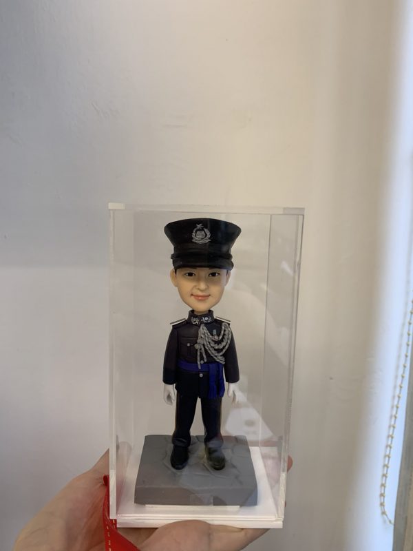 一尊穿著禮服式深色制服的警察人像公仔置於透明展示盒內，由手掌托著展示；公仔頭戴有徽章的制帽，肩章與領章清晰，胸前繫有銀灰色繩結與勳表，腰間藍色飾帶；雙手配白手套、黑色皮鞋，站在方形石磚紋台座上。背景為淺色牆面與窗邊捲簾繩。整體呈現正式典禮感，適合紀念與送禮。
