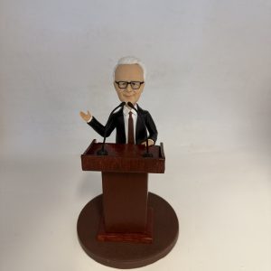 新聞發佈會主持人主題人像公仔：短白髮、戴黑框眼鏡的男士身穿黑色西裝與深色領帶，站在木紋講台後，雙手分別做講解與持講稿姿勢，面前設有兩支黑色麥克風；圓形棕色底座與素色背景突顯正式的發佈會場景。適合作為新聞發佈會主持人人像公仔的榮休禮物與展示擺設。