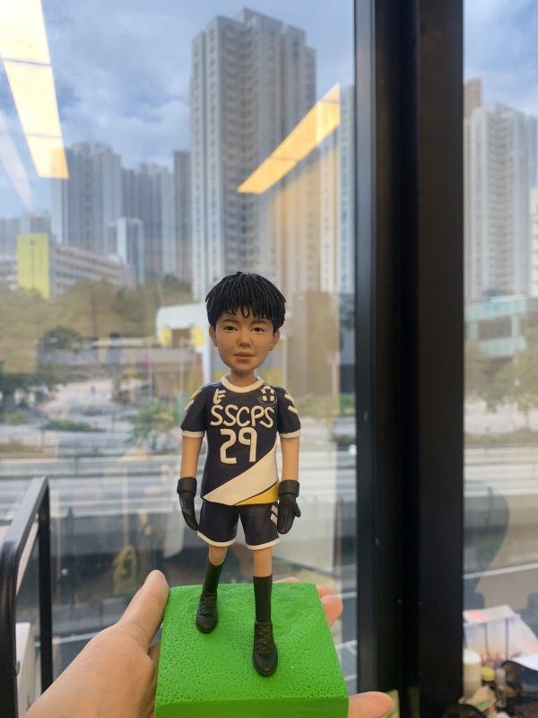 手掌上托著一尊運動主題的人像公仔，穿深藍與白色斜紋設計的球衣與短褲，胸前印有字樣「SSCPS」與號碼「29」，肩位有徽章裝飾；公仔戴黑色運動手套、穿長襪與黑色球鞋，站在亮綠色草地質感方形底座上。背景為落地窗外的城市高樓景觀與道路，室內燈光在玻璃上有反射。整體呈現青春活力與比賽氛圍。Keywords：人像公仔，運動禮物。