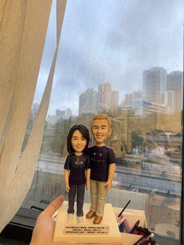 雙人牧師人像公仔：兩位人物並肩貼近、自然微笑，穿深紫團體T恤與休閒長褲；女方配白色運動鞋、男方配棕色皮鞋。立於長方形可刻經文與祝福文字的底座，適合作為教會致謝、授職、周年或生日的人像公仔禮物。