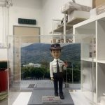 香港警察學員結業禮人像公仔，穿著白色制服襯衣與深色長褲，頭戴黑色警帽，帽上印有紫色徽章，胸前配有名牌與肩章。公仔被安置於透明展示盒內，背景是香港警察訓練學校的山景與操場。底座字牌顯示「Intake 01/2023 結業禮」，象徵完成學員培訓的重要時刻，專為紀念典禮而設計。