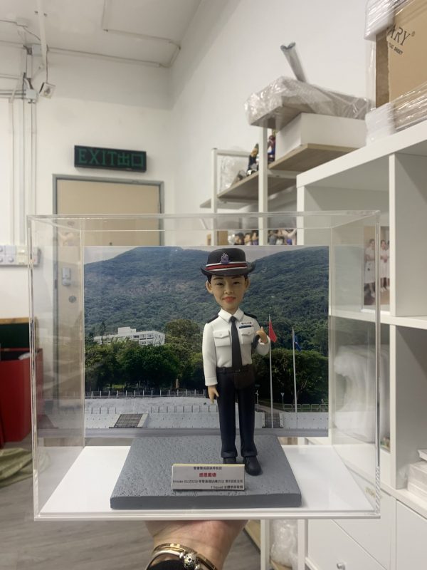香港警察學員結業禮人像公仔，穿著白色制服襯衣與深色長褲，頭戴黑色警帽，帽上印有紫色徽章，胸前配有名牌與肩章。公仔被安置於透明展示盒內，背景是香港警察訓練學校的山景與操場。底座字牌顯示「Intake 01/2023 結業禮」，象徵完成學員培訓的重要時刻，專為紀念典禮而設計。