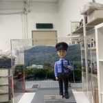 「香港警察學員人像公仔，身穿淺藍色長袖制服襯衫，佩戴黑色肩章與領帶，頭戴警帽，胸前名牌清晰可見，右手自然下垂，左手則做出敬禮般的姿勢，展現莊重與自信。公仔置於透明展示盒中，背景為綠意盎然的山景與訓練場地，象徵警隊訓練與守護社會的使命。底座附有字牌，內容提及『Intake 01/2023 警察學員結業禮』，可見此人像公仔為一份專屬訂製的結業紀念禮物，寓意學員順利完成培訓，邁向警務生涯的新起點。」