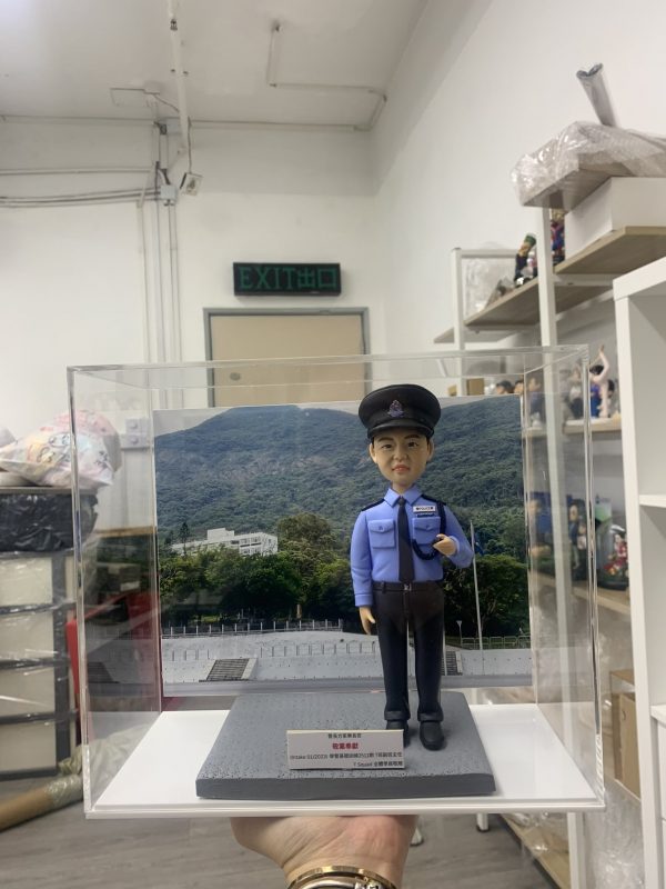 「香港警察學員人像公仔，身穿淺藍色長袖制服襯衫，佩戴黑色肩章與領帶，頭戴警帽，胸前名牌清晰可見，右手自然下垂，左手則做出敬禮般的姿勢，展現莊重與自信。公仔置於透明展示盒中，背景為綠意盎然的山景與訓練場地，象徵警隊訓練與守護社會的使命。底座附有字牌，內容提及『Intake 01/2023 警察學員結業禮』，可見此人像公仔為一份專屬訂製的結業紀念禮物，寓意學員順利完成培訓，邁向警務生涯的新起點。」