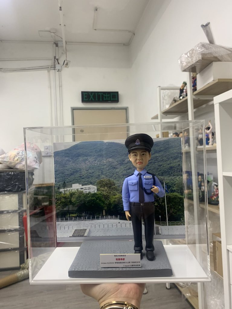 「香港警察學員人像公仔，身穿淺藍色長袖制服襯衫，佩戴黑色肩章與領帶，頭戴警帽，胸前名牌清晰可見，右手自然下垂，左手則做出敬禮般的姿勢，展現莊重與自信。公仔置於透明展示盒中，背景為綠意盎然的山景與訓練場地，象徵警隊訓練與守護社會的使命。底座附有字牌，內容提及『Intake 01/2023 警察學員結業禮』，可見此人像公仔為一份專屬訂製的結業紀念禮物，寓意學員順利完成培訓，邁向警務生涯的新起點。」
