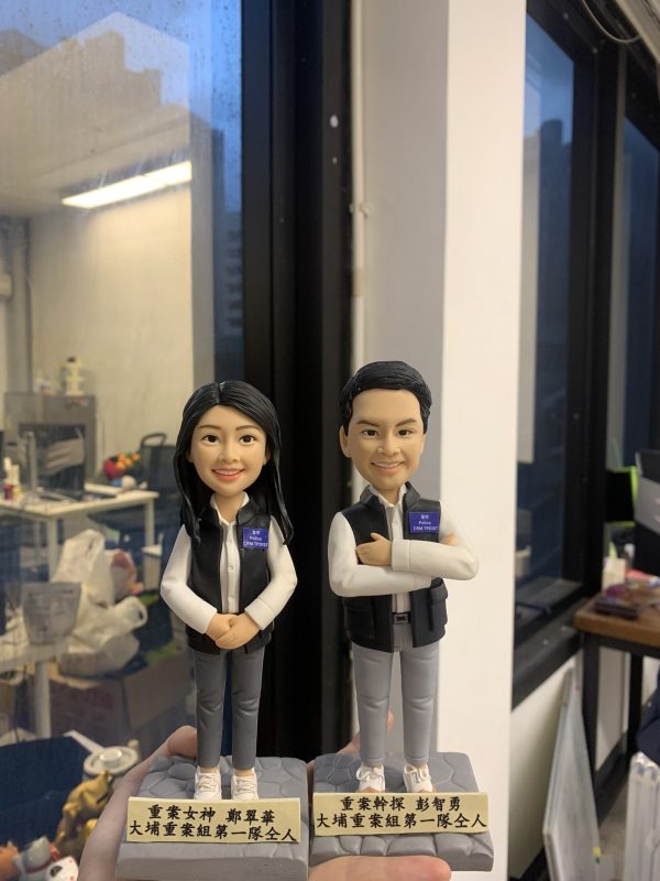 兩尊成對的人像公仔置於透明展示盒外，由手掌襯托拍攝：皆穿白色襯衫、深色無袖背心與淺灰長褲、白色運動鞋。左側為長髮女性公仔，雙手在腹前交握、微笑；右側為短髮男性公仔，右臂環抱左臂呈自信站姿。兩者胸前皆別有深藍識別證牌；腰間可見皮帶與簡化裝備造型。公仔站在石磚紋方形台座，前方金色名牌各以黑字刻有稱呼與致意語。背景是室內工作室環境與窗邊夜色反射，營造致敬與送禮紀念氛圍。