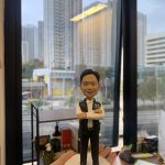 男性警察人像公仔，職級標示為總督察；短髮微笑，白襯衫配深色長褲與黑鞋，外穿深色戰術背心，胸前印有 POLICE 與臂章圖樣；雙臂交叉站姿。公仔立於白色圓形台座，前方金色名牌刻有祝賀升遷與贈送者資訊。背景為室內辦公環境與落地窗外的高樓城市景觀，由手掌托起近距離拍攝，呈現紀念與送禮氛圍。