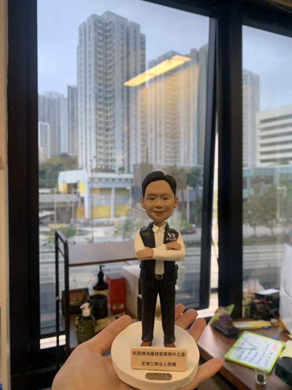 男性警察人像公仔，職級標示為總督察；短髮微笑，白襯衫配深色長褲與黑鞋，外穿深色戰術背心，胸前印有 POLICE 與臂章圖樣；雙臂交叉站姿。公仔立於白色圓形台座，前方金色名牌刻有祝賀升遷與贈送者資訊。背景為室內辦公環境與落地窗外的高樓城市景觀，由手掌托起近距離拍攝，呈現紀念與送禮氛圍。