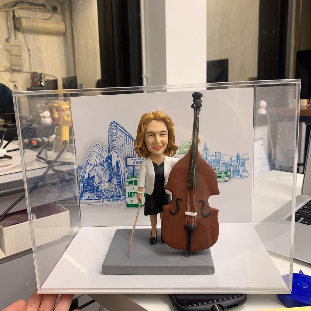 一款為女性低音大提琴演奏家訂製的人像公仔,公仔站在樂器旁,其背景配件裝飾為一張印有香港電車、渡輪及城市景觀的手繪風格圖畫。