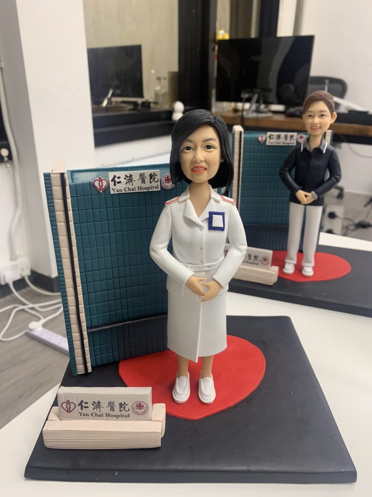 訂製仁濟醫院醫護人像公仔｜身穿白色醫師袍的醫生公仔站立於醫院屏風前，腳下有紅色心型底座，背景標示「仁濟醫院 Yan Chai Hospital」標誌。此公仔專為醫生與醫護人員打造，是獨特的專屬禮物與紀念擺設。