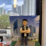 總督察職級主題的人像公仔站在灰色人行道造景台座上，雙臂交叉、短髮微笑；身著白色上衣與深色長褲、藍白運動鞋，外搭黑色戰術背心，胸前可見 POLICE 識別與徽章樣式。公仔背後設有印刷的現代商辦大樓街景背景板；更遠處透過辦公室窗戶可見真實城市高樓與藍天白雲，窗面映出室內燈條反光。前景散見書籍、文具等物件，營造都會與職場氛圍，呈現紀念與送禮用途。