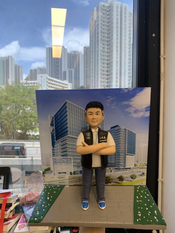 總督察職級主題的人像公仔站在灰色人行道造景台座上，雙臂交叉、短髮微笑；身著白色上衣與深色長褲、藍白運動鞋，外搭黑色戰術背心，胸前可見 POLICE 識別與徽章樣式。公仔背後設有印刷的現代商辦大樓街景背景板；更遠處透過辦公室窗戶可見真實城市高樓與藍天白雲，窗面映出室內燈條反光。前景散見書籍、文具等物件，營造都會與職場氛圍，呈現紀念與送禮用途。