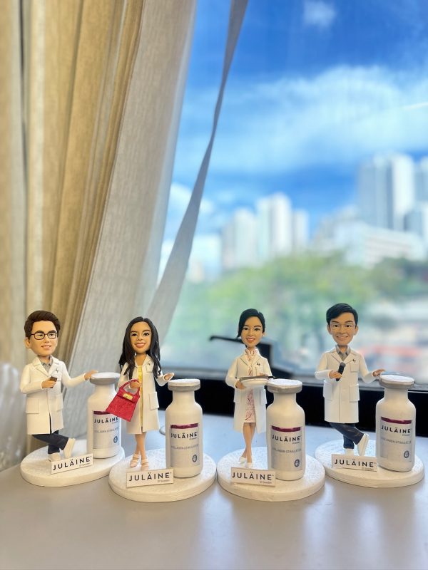 四位穿白袍的醫生／醫護人像公仔排成一列，背景為窗外藍天與高樓景致；每個公仔站在印有 JULAINE 字樣的底座旁，身側設有放大版「膠原增生注射」示範瓶模型，其中兩位手持托盤與疑似注射器，細節精緻，呈現醫學美容臨床感；整體作為醫生、醫護致謝或退休禮物極具紀念性。