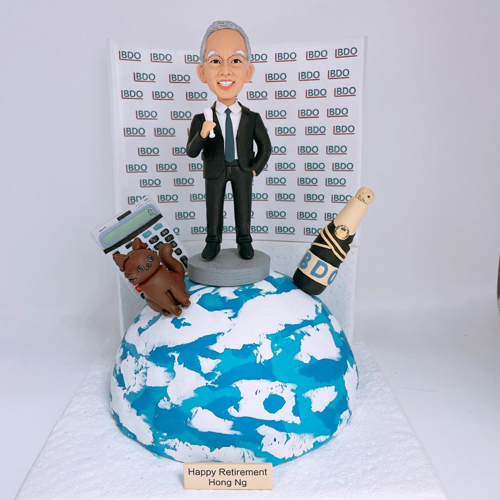 老闆退休主題的人像公仔：銀灰短髮、戴眼鏡的男士身穿黑色西裝與領帶，站在小型圓台上比出讚的手勢；前方為藍白地球造型底座，左側擺放大型計數機與棕色貓咪公仔，右側有綁著黑金緞帶的紅酒香檳瓶並貼有「BDO」字樣；背板重複排列「BDO」標誌，前方銘牌寫有「Happy Retirement」。整體呈現職場成就與個人喜好的退休禮物人像公仔佈置。