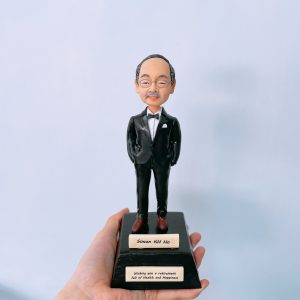 一款為老闆退休訂製的人像公仔,描繪了一位戴眼鏡的年長男士,他身穿黑色燕尾服,站立在一個刻有姓名和退休祝福語的黑色獎盃底座上。