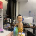 手掌上托著一尊兒童造型的游水人像公仔。公仔留短髮，赤裸上身，穿著黃色印有紅色小魚圖案的泳褲，右手自然下垂，左臂抱著綠藍雙色圓形泳圈。腳踩貝殼與細沙質感的底座，旁邊放著藍黃配色的小蛙鞋與小水池邊緣造型。Keywords：人像公仔，游水人像公仔。