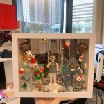 一個放在白色展示盒內的客製化人像公仔，場景以聖誕節為主題，周圍裝飾有聖誕老人、公仔雪人、聖誕樹、馴鹿與聖誕燈飾，背景營造溫馨的節日氛圍，非常適合作為聖誕節禮物或驚喜心意。