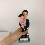 父女合影主題的警察人像公仔作品，男士以短髮造型、穿深色外套配白襯衫，面帶溫和笑容，左臂自然環抱身穿粉紅色長袖連身裙的小女孩，女孩綁著小髻、笑眼彎彎，雙手交疊靠在父親胸前，呈現親密而溫馨的互動；兩人站在四角帶有綠邊與彩色小圖案點綴的方形底座上，前側貼有可客製的姓名與職級名牌（示例文字：YAU Yu-sing｜CIP CRM YTDIST），五官刻畫立體、眉眼與嘴角弧度清晰，頭髮以細束刻紋呈現層次，衣物的翻領、衣襬與皺褶以陰影疊色表現質感，可依要求更換或追加背景與配件如辦公室或指揮室場景、勳章與隊章元素、座駕模型或透明展示盒，適合作為警務人員榮升、榮休、嘉許、周年與生日等多種場合的送禮與收藏之選；關鍵字：警察人像公仔、送禮。