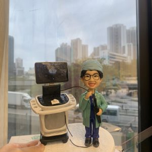 醫生人像公仔，身穿綠色手術袍與藍色醫師服，戴著眼鏡微笑站立，手持內視鏡操作器械。旁邊有超聲波內視鏡診斷儀器與螢幕，場景高度還原臨床醫療環境。此人像公仔是專屬醫生、中醫師的客製化禮物，適合送給醫護人員作為紀念或感謝之用。