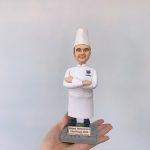 手掌托起的廚師主題人像公仔：戴高帽、穿全白廚師服的男廚師雙臂交叉微笑站立，胸口左側繡有彩色圖案與姓名字樣。腳下是灰色石磚紋底座，前方金色名牌刻著「Happy Retirement Chef Rudy 2023」及贈送者名字。背景為素色牆面，焦點集中在公仔細節與典禮感十足的退休題字，呈現典雅的廚師退休禮物擺設。