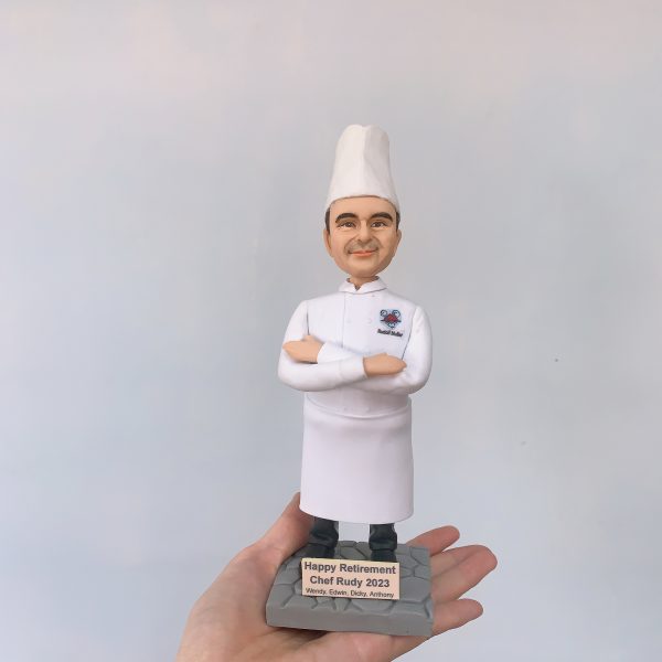 手掌托起的廚師主題人像公仔：戴高帽、穿全白廚師服的男廚師雙臂交叉微笑站立，胸口左側繡有彩色圖案與姓名字樣。腳下是灰色石磚紋底座，前方金色名牌刻著「Happy Retirement Chef Rudy 2023」及贈送者名字。背景為素色牆面，焦點集中在公仔細節與典禮感十足的退休題字，呈現典雅的廚師退休禮物擺設。